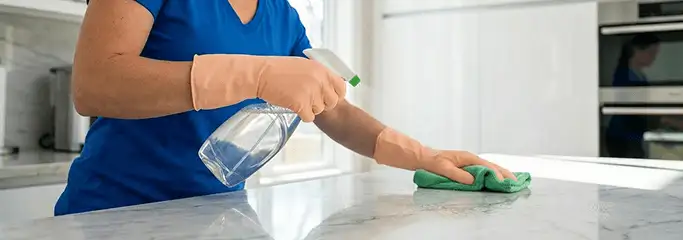 Maid service worker spraying and wiping a kitchen countertop with a microfiber cloth for sanitizing, Dallas, TX.