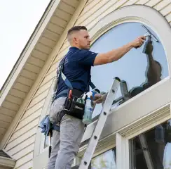 Professional window cleaner on a ladder using a squeegee to clean an exterior arched window, Dallas, TX.
